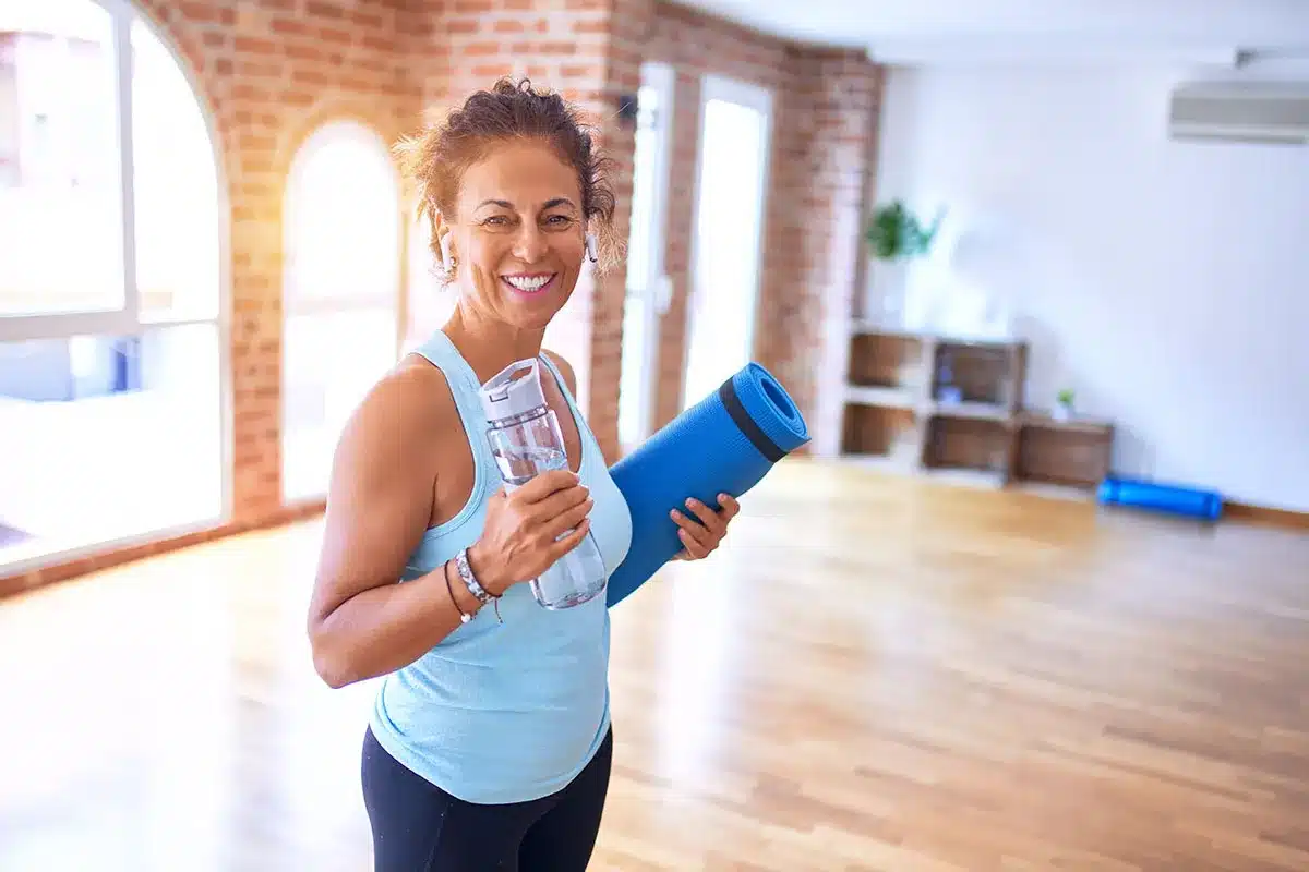 Smiling woman holding a water bottle and yoga mat.