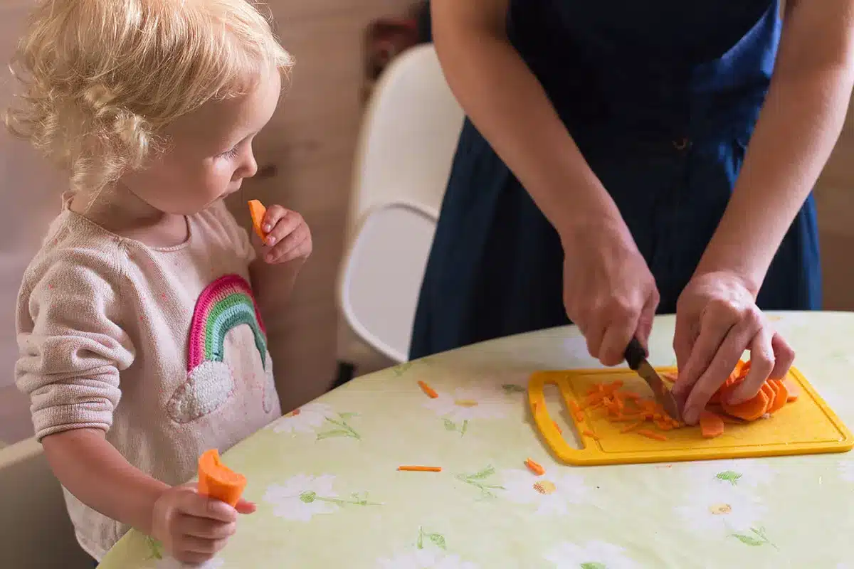 Toddler eats carrot while mom prepares food.