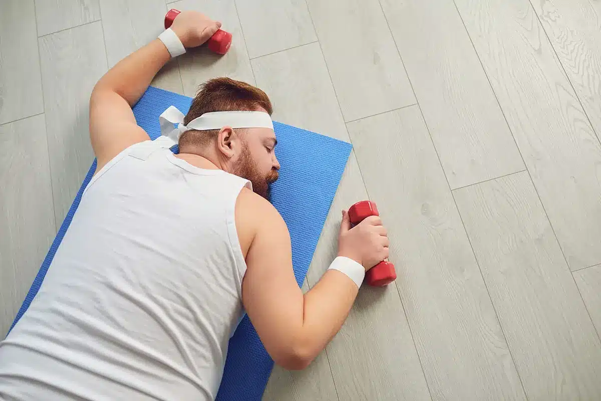 Overweight man sleeping on yoga mat with dumbbells.
