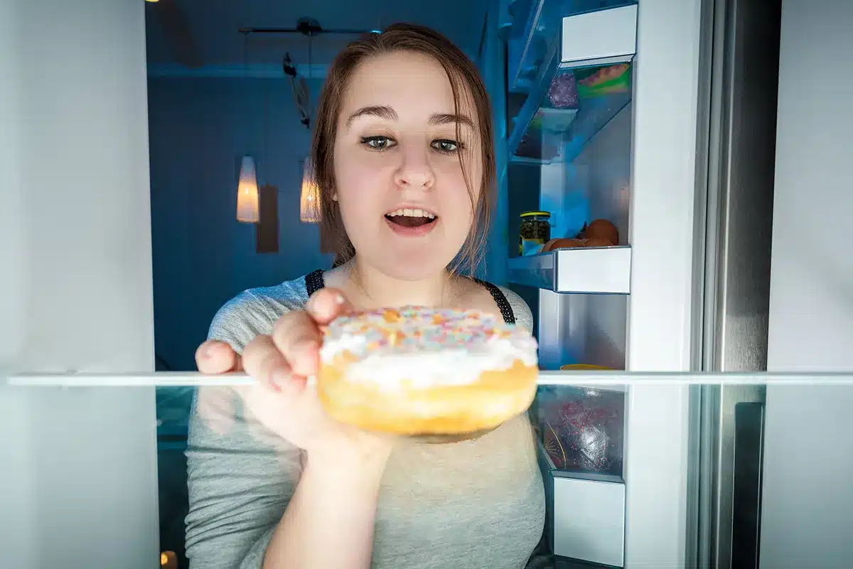 Woman reaching for donut in refrigerator at night.