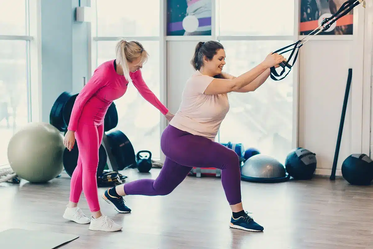 Plus-size woman doing TRX lunges with trainer.