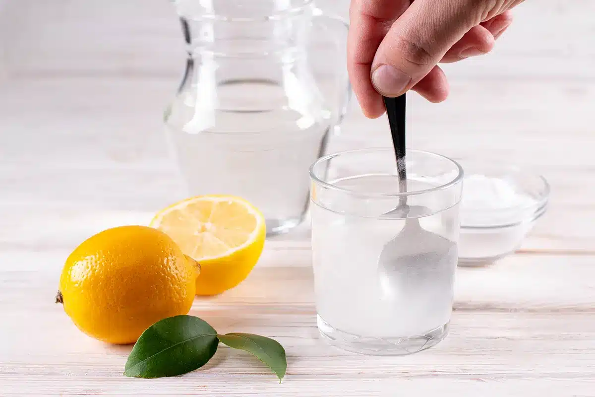 A hand adds baking soda to water in a glass. Lemons and a bowl of baking soda are nearby.