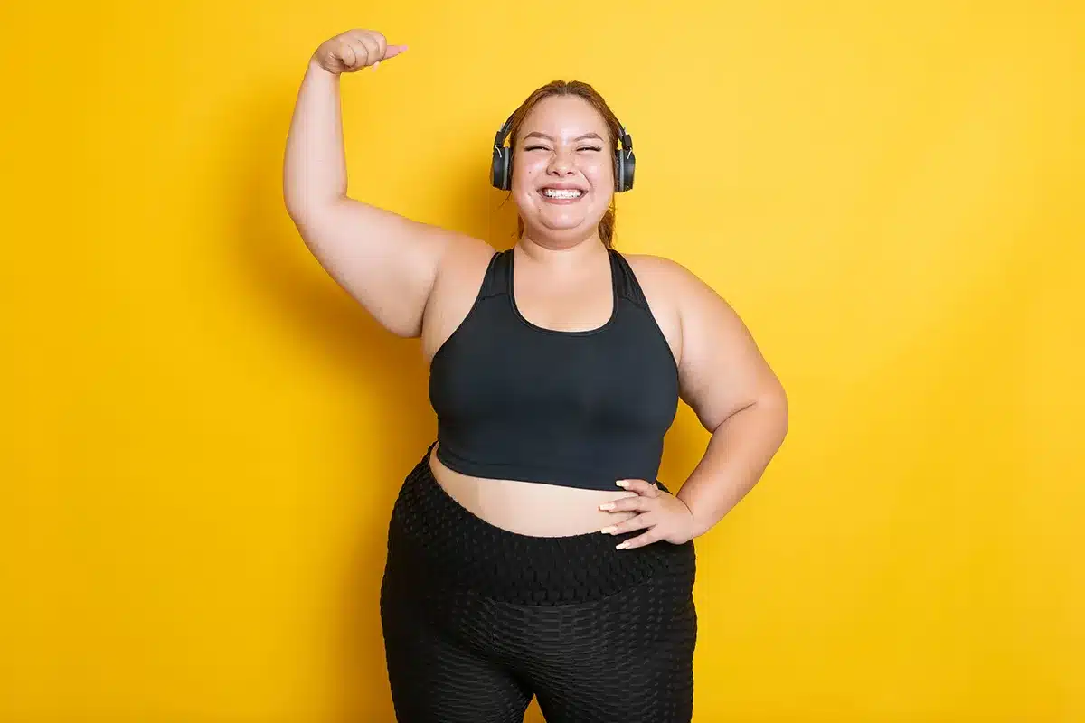 Happy plus-size woman wearing workout clothes and headphones, flexing her bicep.