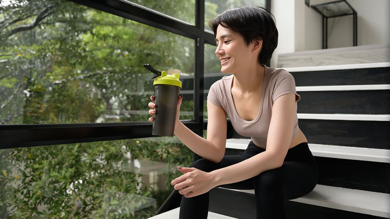 Smiling woman in athletic wear holding a shaker bottle by a window.