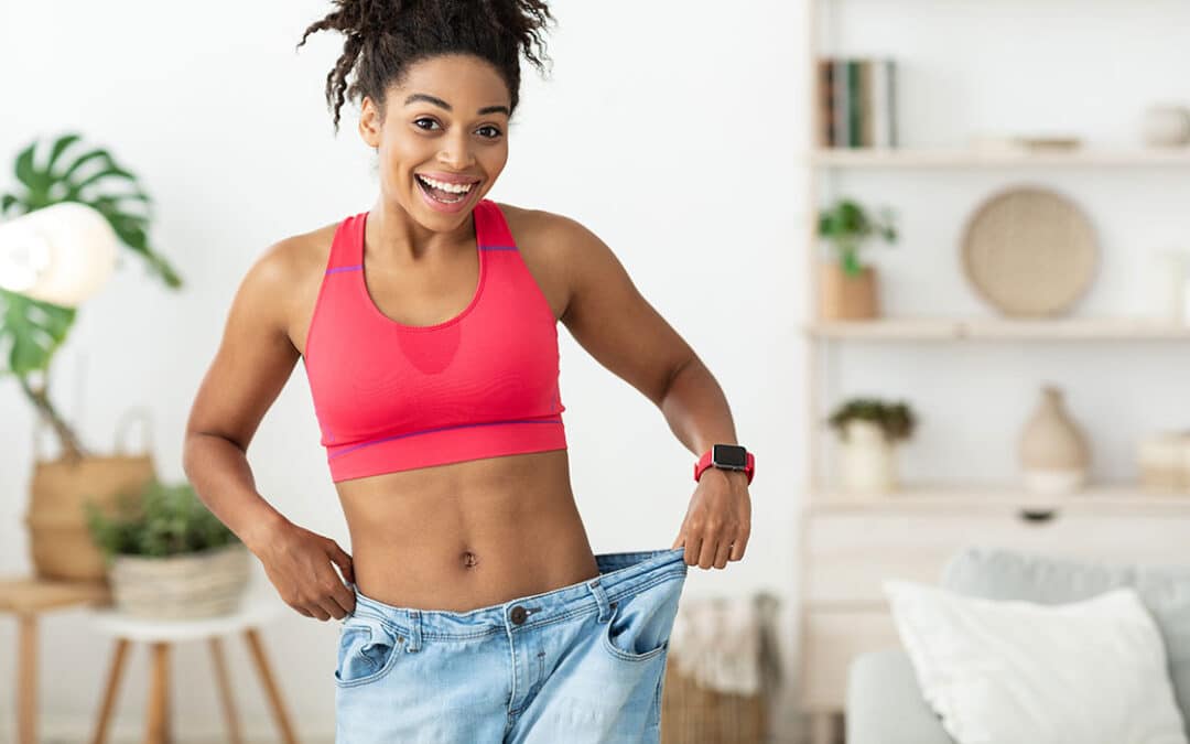 A smiling, fit woman in a sports bra celebrating her body transformation in her old, baggy pants.