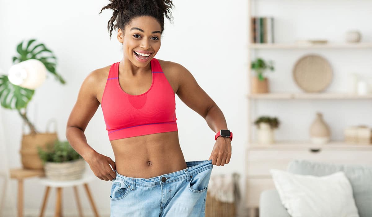 A smiling, fit woman in a sports bra celebrating her body transformation in her old, baggy pants.