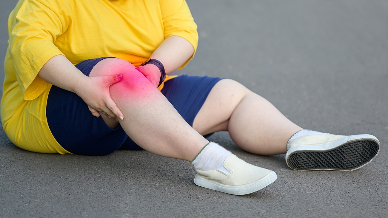 Overweight person sitting on the ground, clutching a knee with highlighted pain.